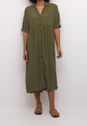 Robe chemise vert olive avec un devant à boutons, des manches courtes retroussées et une forme évasée, fabriquée en tissu léger. Des sandales plates complètent le look.