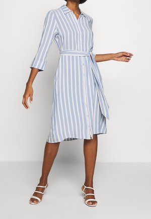 Mujer vestida con vestido camisero de rayas verticales azules y blancas hasta la rodilla, con cinturón en la cintura y sandalias blancas de tacón con tiras.