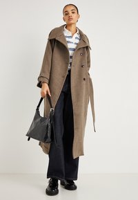 Casaco de lã castanho com gola larga e botões abotoados cruzados, combinado com camisa às riscas, jeans escuros, mala preta e sapatos pretos brilhantes.