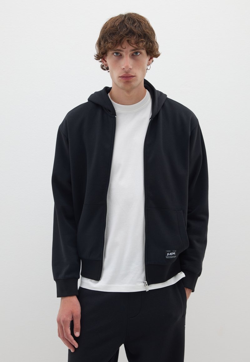 Haut de veste à zip noir avec une poche à l'avant, fabriqué en tissu doux. Il présente une capuche, des poignets côtelés et un petit logo sur l'ourlet. T-shirt blanc en dessous.