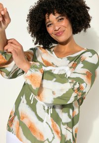 Maglietta a maniche lunghe verde e bianca con un motivo astratto e dettagli arancioni, caratterizzata da un collo a V e maniche arricciate.