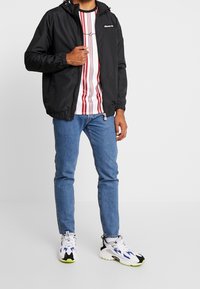 Sort lynlåsfri jakke med hætte, båret over en hvid skjorte med lodrette røde striber, kombineret med blå jeans og flerfarvede sneakers.