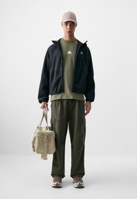 Casaco de fleece preto com fecho, t-shirt verde oliva com logótipo, calças cargo em verde oliva, saco bege e ténis castanhos com pormenores brancos. Fundo simples.