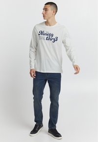 Hvid langærmet skjorte med grafisk tekst i marineblå, parret med blå denim jeans og sorte sneakers. Ren design, afslappet pasform.