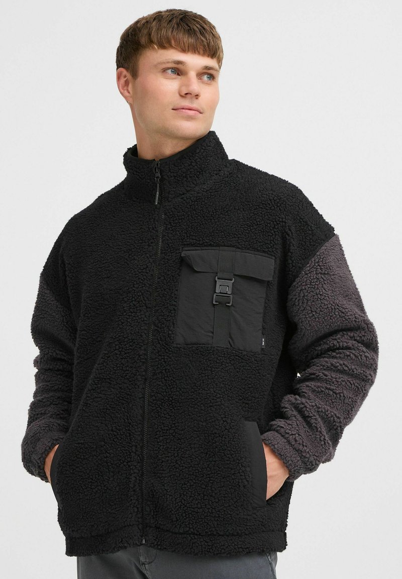 Chaqueta de forro polar negra con cuello alto, tela texturizada, un bolsillo frontal con hebilla y mangas grises en contraste. Cierre de cremallera.