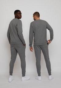 Deux mannequins portent des sweatshirts gris à manches longues assortis et des joggings ajustés, tous deux en matière douce et texturée. Des baskets blanches.