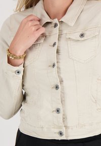 Vrouw in een beige knopenjack, met haar linkervinger de kraag vasthoudend, en met een gouden kralenarmband om haar pols.