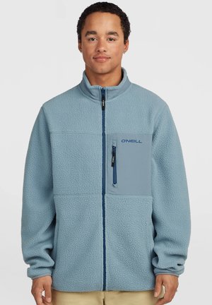 Junger Mann trägt hellblaue Fleecejacke mit Reißverschluss vorne und Brusttasche, steht vor einem einfarbigen hellen Hintergrund.