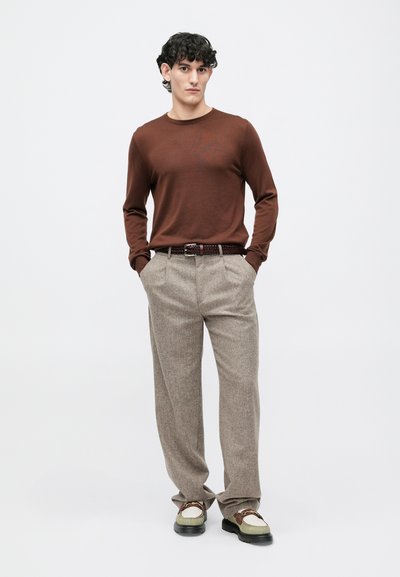 Camisa de manga longa castanha, tecido texturizado, combinada com calças cinza à padrão xadrez, cinto de pele e sapatos em várias tonalidades, de pé contra um fundo branco.