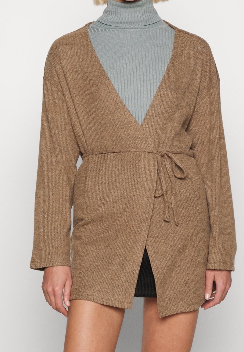 Femme portant un cardigan beige tricoté et noué à la taille, par-dessus un col roulé côtelé bleu clair et une jupe noire.
