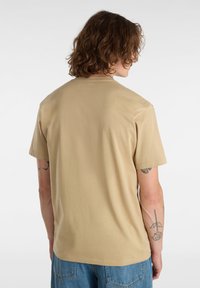 Béžové bavlněné tričko s volným střihem, krátkými rukávy a hladkým zadním dílem, doplněné modrými džínami. Žádné viditelné logo ani vzory.