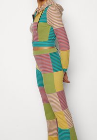 Une combinaison multicolore avec un hoodie court à fermeture éclair. Présente des motifs rayés et en patchwork dans des tons de vert, jaune, violet et bleu.