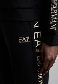 Črna sweatshirt in hlače z zlatim logotipom "EA7 Emporio Armani". Tkanina je gladka in raztegljiva, s sproščenim krojem.