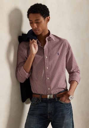 Young man in red checkered shirt with rolled sleeves, dark jeans, and brown belt holds a dark jacket over his shoulder against a plain wall.