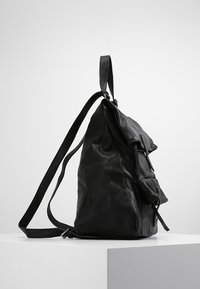 Černý kožený batoh s texturovaným povrchem, nastavitelným popruhem a přední kapsou s klopou. Boční profil.