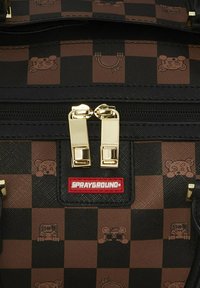 Mochila a cuadros con cuadrados marrones y negros, cremalleras de hardware dorado y diseños de osos de dibujos animados en relieve. Parche de logo rojo visible.