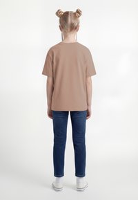 Bērns ar divām matu bulciņām un bizēm, valkājot vienkāršu brūnu T-kreklu, zili džinsus, baltas zeķes un baltas sporta kurpes, stāvot muguru pavērsts.