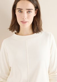 White, textured fabric top with a round neckline and three-quarter sleeves. Features a central seam detail for added design.