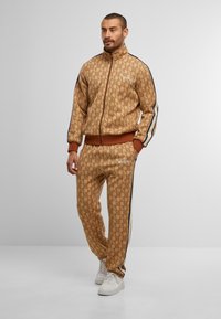 Beanie, zip-up jacket, and pants set in a beige pattern, featuring brown accents and side stripes, made from a smooth, textured fabric.