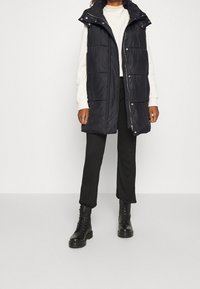 Schwarze Daunenweste mit hohem Kragen, Druckknöpfen und zwei Fronttaschen. Getragen über einem cremefarbenen Pullover und schwarzen Ripp-Hosen mit schwarzen Stiefeln.