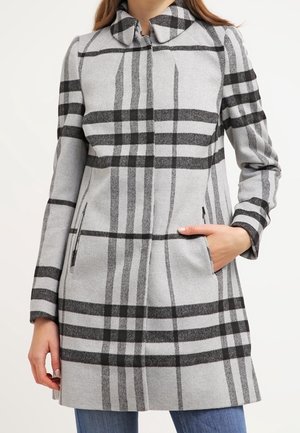 Femme portant un long manteau gris avec un grand motif à carreaux noirs, debout avec une main dans la poche et un jean bleu visible.