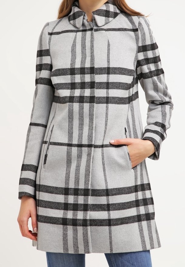 Femme portant un long manteau gris avec un grand motif à carreaux noirs, debout avec une main dans la poche et un jean bleu visible.