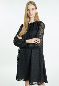 Musta mekko, jossa on teksturoitu läpinäkyvä päällinen ruutukuviolla, pitkät puffihihat ja rento malli, jossa on pyöreä pääntie.