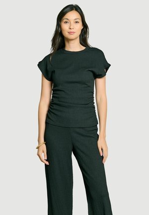 Femme debout portant un haut texturé noir à manches courtes et un pantalon large assorti avec un bracelet beige à son poignet gauche.