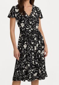 Černé květinové šaty s krátkým rukávem, které mají bílý květinový vzor, výstřih do V a zavazovací pásek v pase. Měkká, splývavá látka.