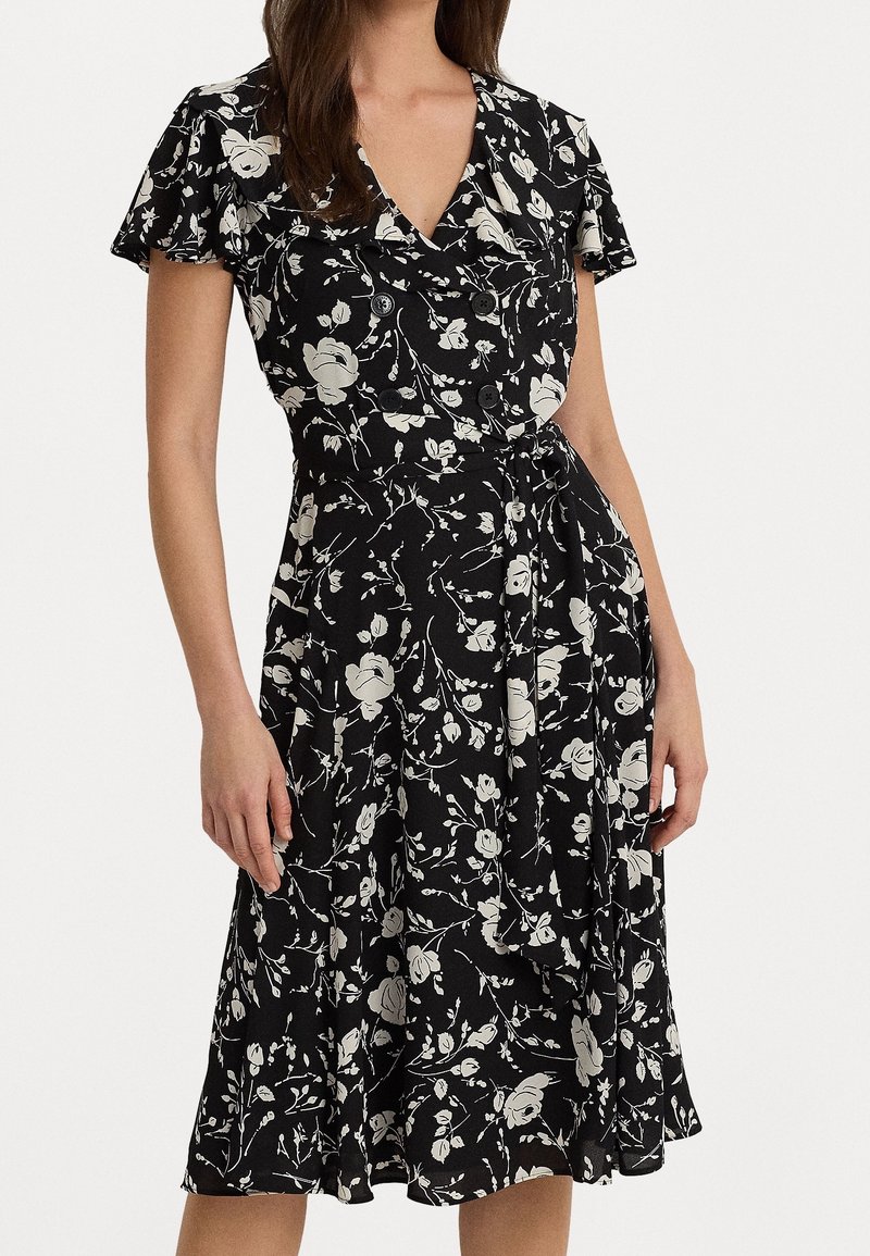 Černé květinové šaty s krátkým rukávem, které mají bílý květinový vzor, výstřih do V a zavazovací pásek v pase. Měkká, splývavá látka.