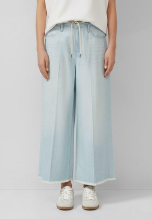 Osoba ubrana w jasnoniebieskie szerokie, skrócone jeansy z postrzępionymi brzegami i białe trampki, stojąca na tle jednolitego szarego tła.