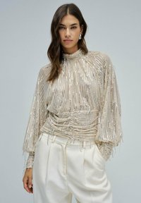 Frau in einer transparenten beigen Bluse mit silbernen Pailletten und hohem Kragen, kombiniert mit hochtaillierten weißen Hosen, vor einem grauen Hintergrund stehend.