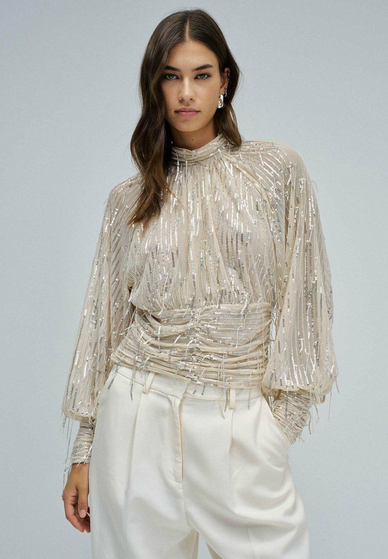 Frau in einer transparenten beigen Bluse mit silbernen Pailletten und hohem Kragen, kombiniert mit hochtaillierten weißen Hosen, vor einem grauen Hintergrund stehend.