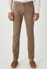 Chinos de algodón marrón con corte slim, cuatro bolsillos y costuras visibles, combinados con zapatos estilo mocasín en color marrón claro.