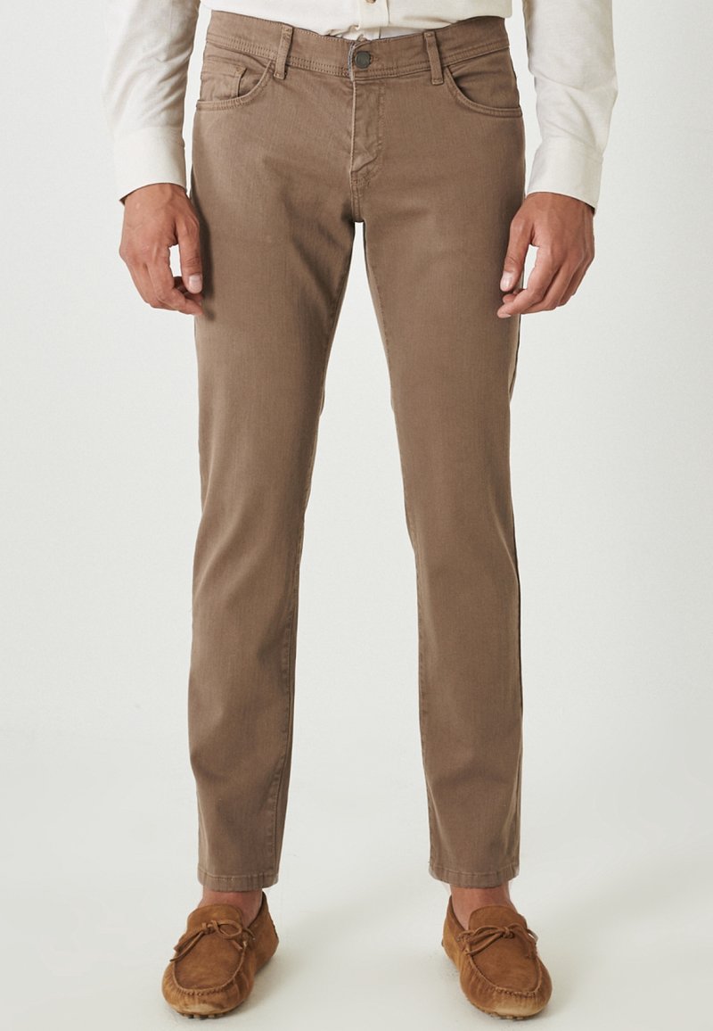 Chinos de algodón marrón con corte slim, cuatro bolsillos y costuras visibles, combinados con zapatos estilo mocasín en color marrón claro.