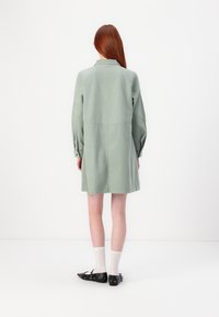 Abito camicia verde oversize e a maniche lunghe, realizzato in tessuto morbido, con colletto e due bottoni sui polsini. Design semplice, lunghezza fino al ginocchio.
