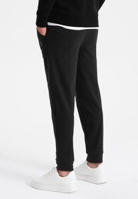 Schwarze Jogginghosen mit schmalem Schnitt, aus weichem Stoff gefertigt. Mit seitlichen Taschen und gerippten Bündchen, kombiniert mit weißen Turnschuhen.