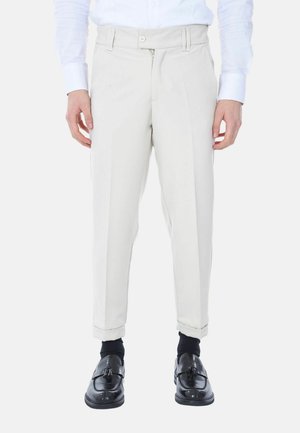 Pantaloni beige chiari con vestibilità slim, dotati di pieghe frontali, chiusura con zip e bottone. Indossati con scarpe nere e calzini.