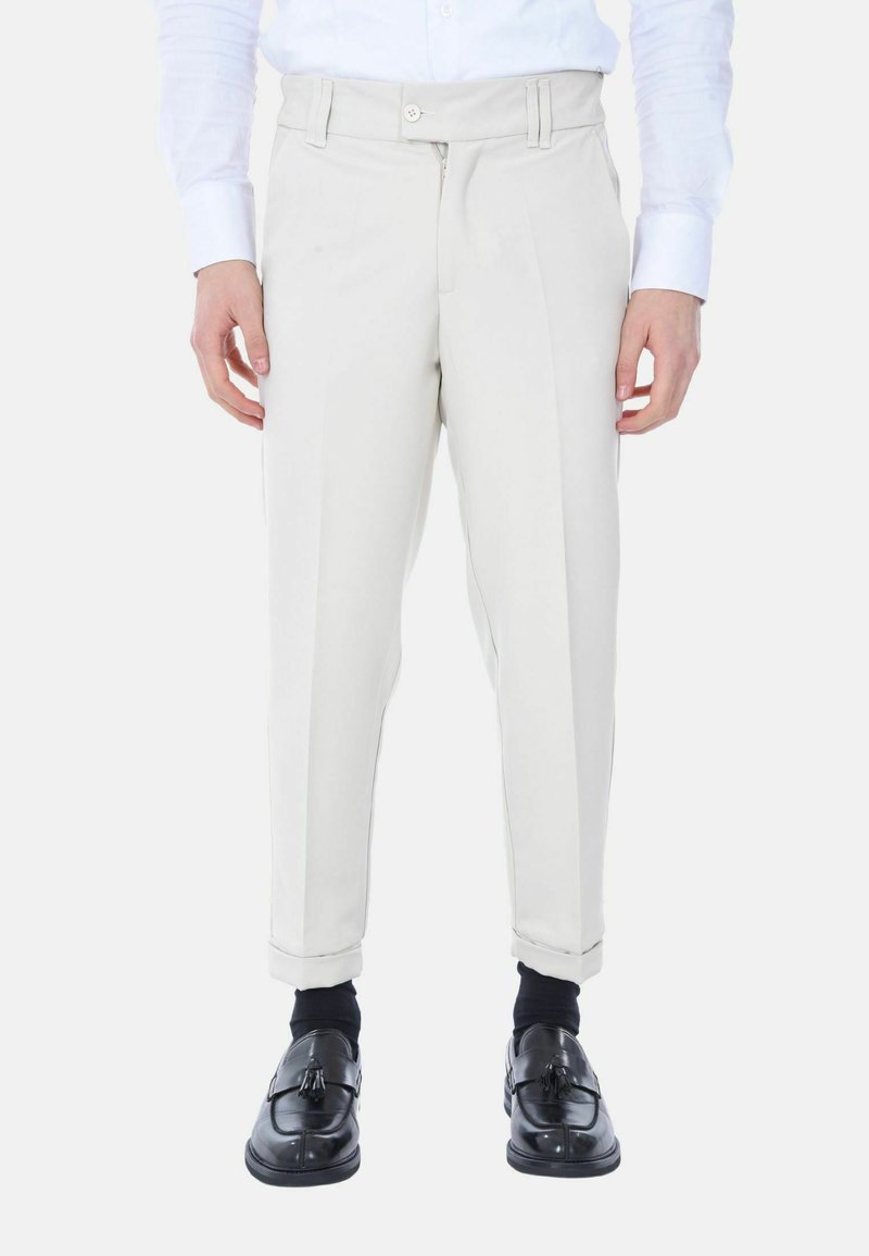 Pantaloni beige chiari con vestibilità slim, dotati di pieghe frontali, chiusura con zip e bottone. Indossati con scarpe nere e calzini.