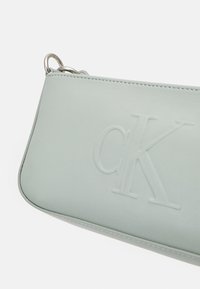 Bracelet en cuir gris clair avec logo "cK" embossé et anneaux métalliques argentés dans le coin supérieur pour attacher la sangle.