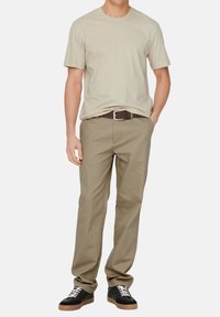 Beige Baumwoll-T-Shirt kombiniert mit khakifarbenen Hosen und schwarzen Sneakern mit Gummi-Sohlen. Das Outfit hat einen braunen Gürtel mit einer Metallschnalle.