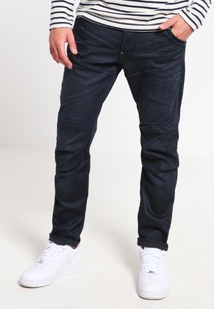 Hombre con pantalones oscuros de corte ajustado, zapatillas blancas y una camisa de manga larga a rayas con las manos en los bolsillos.