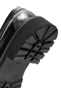 Zapato de cuero negro con suela de goma gruesa que presenta profundas huellas y detalles texturizados, diseñado para durabilidad y tracción.