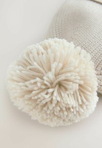 Nahaufnahme einer beigen Strickmütze mit einem großen, flauschigen weißen Wollpompon oben vor einem hellen Hintergrund.