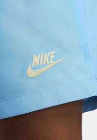 Ljusblått tyg med vit broderad Nike-logotyp och swoosh vid kanten, delvis täckande en persons mörka hudben.