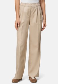 Beige Plissee-Hosen mit geradem Bein, ausgestattet mit genähten Details und einem glatten Stoff. Kombiniert mit braunen Sneakers.