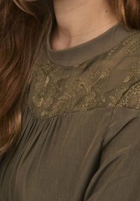 Blouse vert olive avec broderies florales au niveau de l'encolure, tissu texturé, design plissé en dessous du empiècement et col rond.