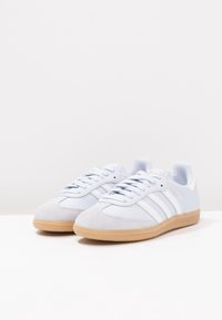 Světlé modré tenisky s horní částí z semiše a kůže, se třemi bílými pruhy a podrážkou z gumy barvy žvýkačky. Mají polstrovaný lem a tkaničky.