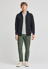 Svart zip-up hoodie, grå t-shirt under, gröna byxor och beige sneakers. Hoodien har framfickor och en subtil logotyp på bröstet.