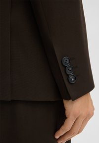 Mouw van een bruin colbert met een gestructureerde stof, voorzien van drie zwarte knopen en stikseldetails nabij de manchetten.